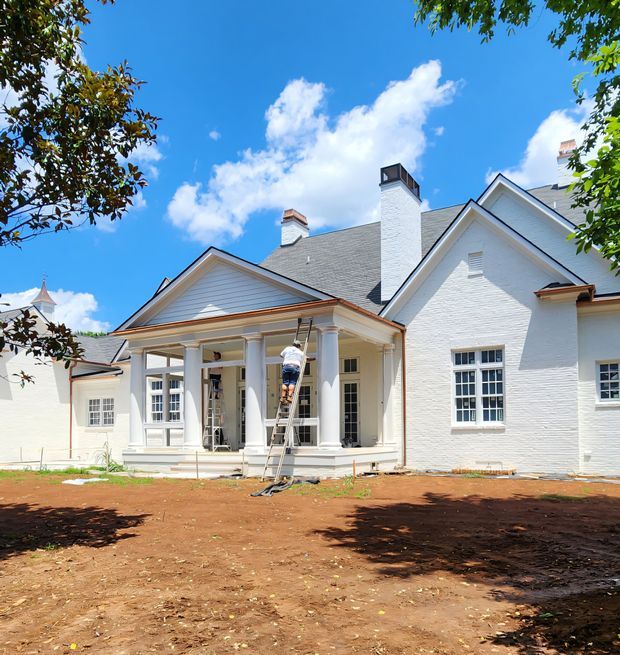 Exterior view of a large white-painted brick residence with prominent gabled roof forms, grey shingle roofing, multiple chimneys, and a covered columned portico. Multi-pane windows and pale trim reinforce a traditional, formal character. The surrounding ground is unfinished earth, suggesting ongoing construction or renovation, with a worker on a ladder at the portico.