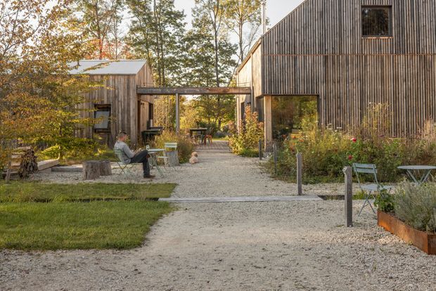 Exterior view of a contemporary timber-clad compound arranged around a landscaped gravel pedestrian courtyard. Two simple gabled buildings frame the central path and are linked by a covered timber walkway. The scene includes planted garden beds, lawn, outdoor seating, raised planters, and mature deciduous trees, creating a warm, informal residential or hospitality setting.