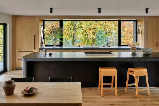 A contemporary open-plan kitchen and dining area with a large dark island, integrated sink and cooktop, warm timber cabinetry, and broad black-framed windows overlooking autumn foliage. Two timber bar stools sit at the island, while a light timber dining table occupies the foreground. Minimal black ceiling spotlights and simple natural decor complete the calm, refined interior.