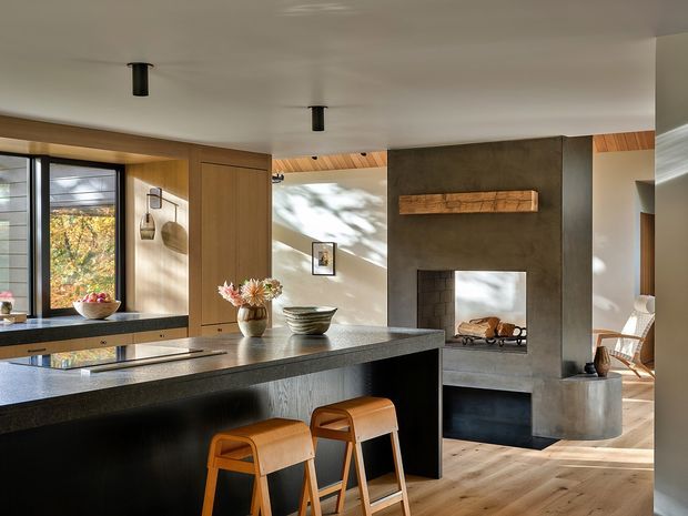A warm, open-plan kitchen and living area with a large dark stone island and two timber bar stools in the foreground. Floor-to-ceiling glazing and timber-framed cabinetry line the left side, while a substantial dark plaster or concrete fireplace with a timber mantel anchors the adjoining living space. Timber flooring, exposed timber ceiling lining, restrained furnishings, and soft natural light create a refined contemporary interior.