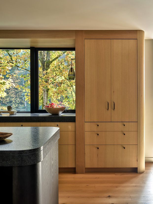 A warm, refined kitchen interior featuring extensive light-to-mid timber cabinetry with rounded vertical trim, integrated drawers, and black handles. A dark stone island benchtop with softened corners occupies the foreground, while black-framed windows overlook leafy trees. Timber flooring, a dark window sill, and understated pendant lighting reinforce the natural, contemporary design.