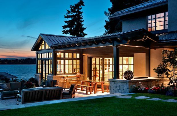 Twilight exterior view of a substantial lakeside home with a glazed timber-framed extension, covered outdoor entertaining area, dark metal pitched roofs, stone-clad base, and illuminated interior spaces. A furnished patio and manicured lawn occupy the foreground, with the lake visible beyond.