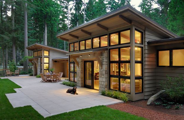 Exterior view of a contemporary single-storey house set among dense trees. The home features dark horizontal cladding, extensive black-framed glazing, exposed timber eaves and beams, and a stone-clad entry volume. A paved patio and outdoor dining area connect the main house with a smaller adjoining structure.