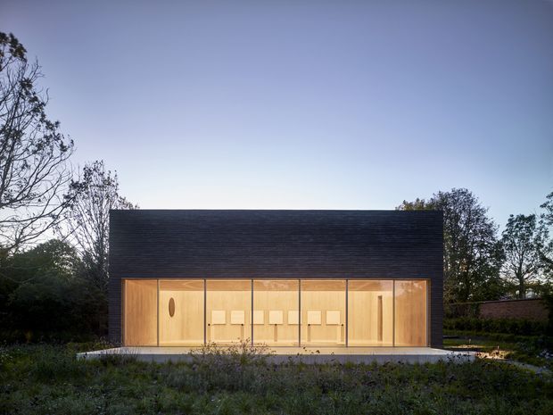 A compact, single-storey modern pavilion set within a landscaped garden. The building has a simple rectangular form, a broad dark pitched roof, and a fully glazed front facade with warm timber-framed openings and illuminated timber interior joinery. Vegetation surrounds the structure, creating a quiet, contemporary garden setting.
