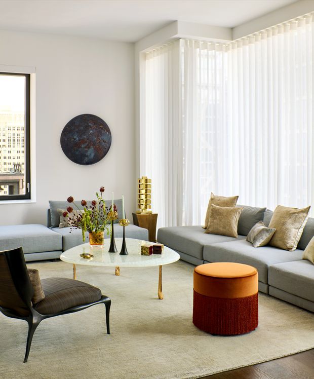 Bright contemporary living room with expansive floor-to-ceiling corner glazing dressed in sheer white curtains. A large pale grey sectional sofa surrounds a round white coffee table, with a sculptural black lounge chair, textured neutral cushions, a large light rug, and a cylindrical orange and deep red upholstered ottoman. Minimal white walls, polished metal accents, and a circular dark artwork create a refined, airy interior.