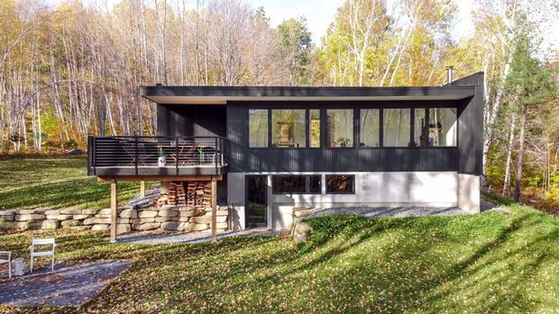 Exterior view of a compact modern multi-level house set within a wooded landscape. The dark upper volume has extensive horizontal glazing, a projecting flat roof, and a deck with black horizontal railings. A stone retaining wall supports the lower level, which combines pale concrete or rendered surfaces with dark-framed windows.