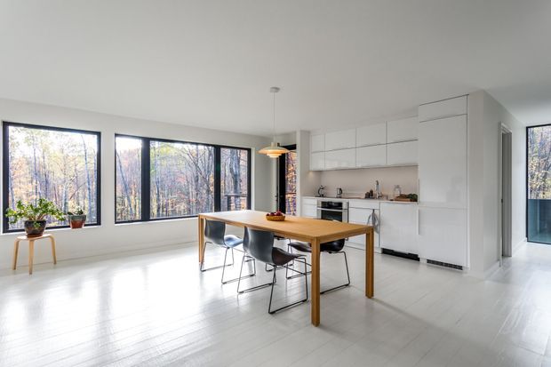 A bright, open-plan dining and kitchen area with a minimalist white interior. A timber dining table with contemporary chairs sits beneath a warm pendant light, while a streamlined white kitchen with integrated cabinetry and appliances occupies the rear wall. Large dark-framed windows provide views of trees and abundant natural light. The room has pale timber or painted flooring and a clean, airy atmosphere.