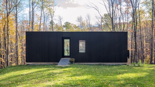 A compact, single-storey rectangular dwelling set within a wooded landscape. The building has a minimalist dark facade with vertical cladding, a flat roof, a narrow glazed entry door, and a small vertical window. A simple concrete path leads across the lawn to the entrance, with deciduous forest surrounding the home.