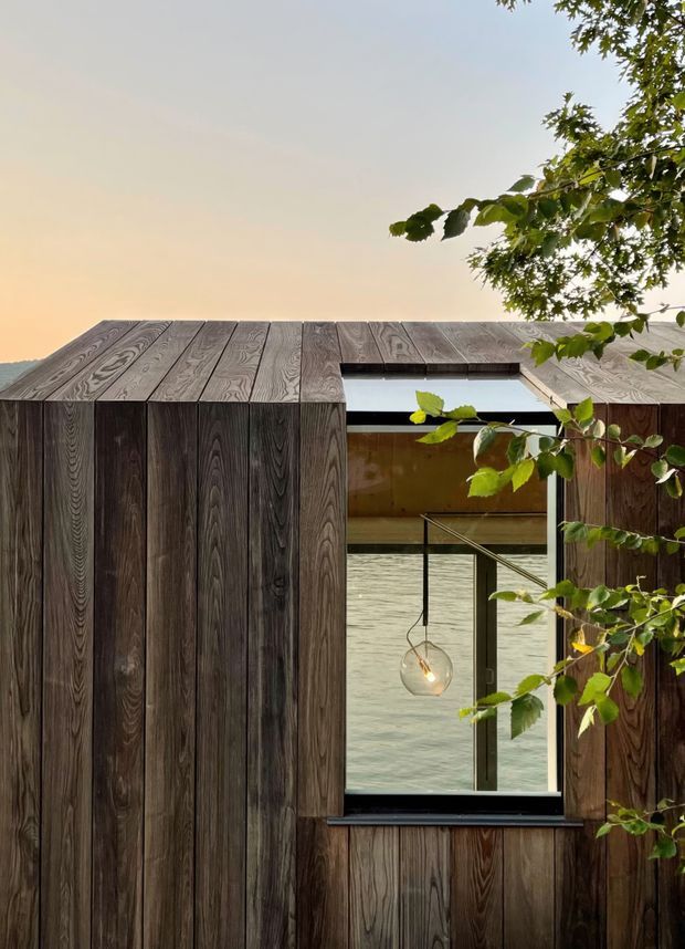 A close exterior view of a compact, sculptural building clad in dark vertically arranged timber boards, with the same timber continuing across the shallow pitched roof. A large recessed window or glazed opening reveals a warm timber interior, pendant light, and water beyond. Tree branches frame the upper and right edges, creating a secluded waterfront setting.