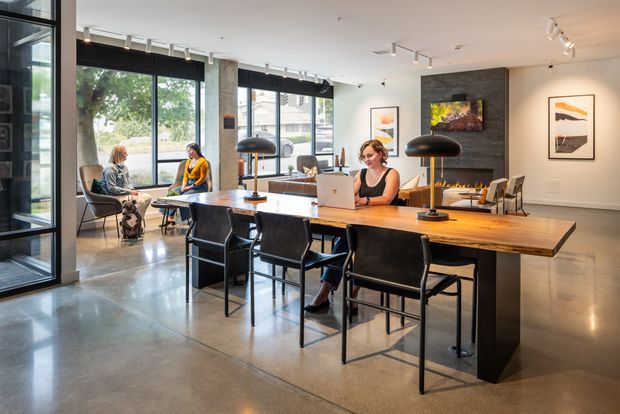 Open-plan contemporary office or shared workspace with a long timber communal table, black chairs, task lamps, and a woman working on a laptop. The space features polished concrete flooring, large black-framed windows, exposed concrete columns, white ceilings with track lighting, and a dark stone fireplace wall with integrated shelving and artwork. Additional seating is arranged near the glazing.