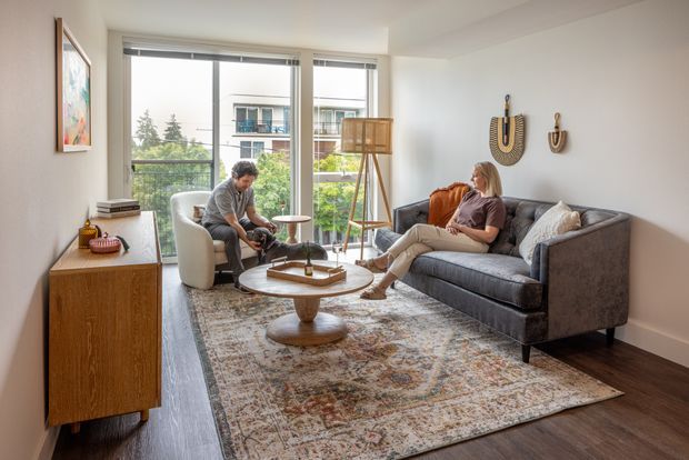 Bright contemporary living room with floor-to-ceiling glazing overlooking greenery and neighbouring apartment buildings. The room features a dark grey sofa, sculptural wood coffee table, patterned area rug, light upholstered lounge chair, timber sideboard, tripod floor lamp, and neutral walls with decorative wall hangings.