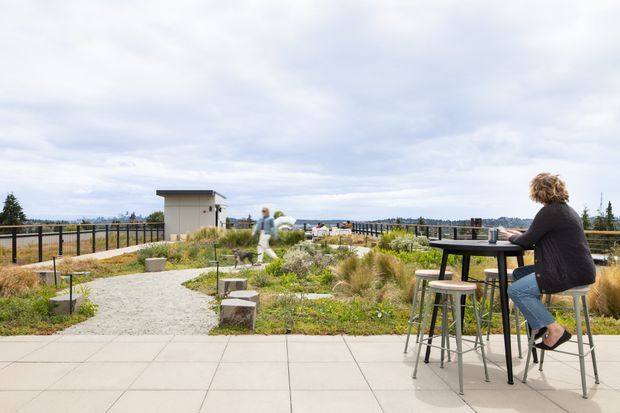 A rooftop terrace or elevated outdoor communal space with paved and gravel areas, landscaped planting beds, black metal railings, and casual high bar seating. A person sits at a tall outdoor table, while another person walks through the garden area. The scene has an open sky, distant landscape views, and a relaxed contemporary rooftop-garden atmosphere.