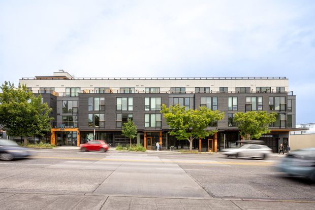 Street-facing view of a contemporary multi-storey apartment or mixed-use building. The facade combines dark charcoal cladding with light horizontal bands, extensive rectangular glazing, balconies, and a flat roofline. Ground-level commercial or lobby entrances open onto a broad urban street lined with small trees, with motion-blurred traffic in the foreground.