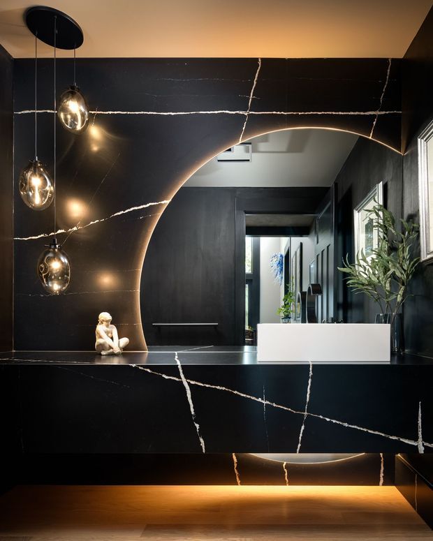 A dramatic, dark-toned residential entryway or hallway featuring a sweeping arched opening clad in black stone with prominent white veining. Three suspended globe pendant lights hang at the left, while a long black stone console or partition with integrated warm underlighting spans the foreground. A white rectangular planter with greenery sits on the counter, and a corridor with dark joinery and reflective surfaces extends beyond the arch.