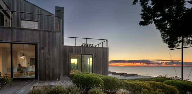 A contemporary multi-level coastal residence clad in weathered vertical timber, photographed at dusk beside calm water. Large glazed openings reveal a warmly lit dining area, while a rooftop terrace with slim metal balustrading overlooks the shoreline. Low native landscaping, decking, and a simple outdoor swing frame the waterfront setting.