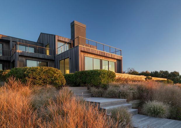 A contemporary multi-level residential house set into a landscaped coastal-looking site, photographed at warm sunset light. The dark timber-clad and glazed facade features projecting volumes, balconies with slim metal balustrades, a prominent brick chimney, and a rooftop terrace. Timber steps and paving lead through ornamental grasses and clipped shrubs toward the home.