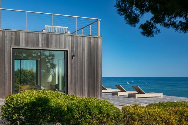 A modern coastal residence viewed from the landscaped exterior, clad in dark vertical timber with a rooftop terrace enclosed by a slim metal balustrade. Large glazed sliding doors reveal a furnished interior, while a timber deck with sun loungers overlooks the ocean. Low clipped shrubs frame the foreground and the scene is brightly lit under a clear blue sky.
