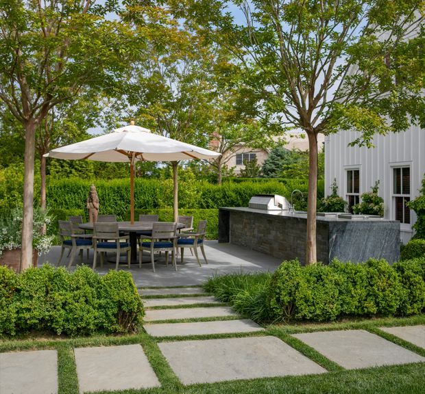 Landscaped backyard entertaining area with a paved patio, outdoor dining table beneath a large white umbrella, built-in stone outdoor kitchen or barbecue counter, manicured hedges, mature trees, and a contemporary light-coloured house facade.