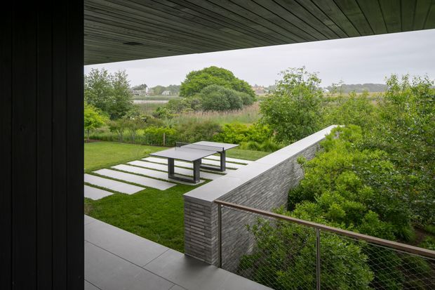 A covered outdoor terrace overlooks a landscaped garden and waterway beyond. The contemporary space features a dark timber-lined soffit, large grey paving slabs, a low grey masonry privacy wall, and a slim metal-and-glass balustrade. A minimalist outdoor dining table sits on a lawn with rectangular stepping slabs, surrounded by dense greenery and mature trees.