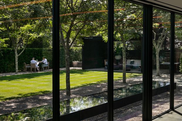 View from inside a highly glazed modern building toward a landscaped courtyard garden. Floor-to-ceiling black-framed glazing divides the foreground, reflecting the garden and a narrow reflecting pool. Outside is a manicured lawn, gravel paths, clipped hedges, mature trees, outdoor seating, and a dark rectangular garden structure. The scene has a refined, tranquil contemporary character.