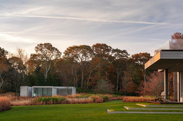 Contemporary modern house exterior at the edge of a landscaped lawn and woodland, photographed in warm autumn light. The visible building has a crisp flat roof, dark horizontal fascia, extensive glazing, and a covered terrace or entry supported by slender columns. Ornamental grasses, layered planting, and a small detached glass-and-metal pavilion complement the minimalist architecture.