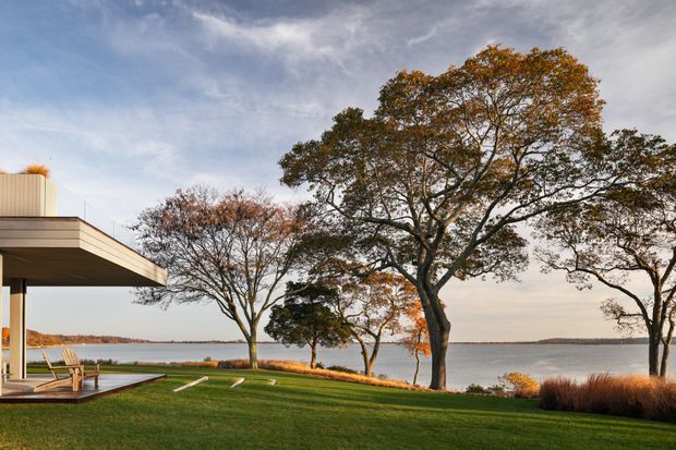 A contemporary waterfront residence viewed from the landscaped grounds, with a broad flat-roofed overhang and glazed living area at left. A manicured lawn slopes toward a calm body of water, framed by mature deciduous trees and ornamental grasses in an autumnal setting.