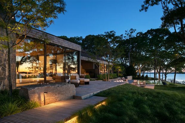 Twilight exterior view of a low-slung contemporary residence set within landscaped grounds beside a body of water. The home features extensive floor-to-ceiling glazing, dark structural framing, a flat roof, stone-clad walls, timber decking and illuminated pathways. Indoor living areas are visible through the glass, while mature trees and waterfront seating create a secluded outdoor setting.