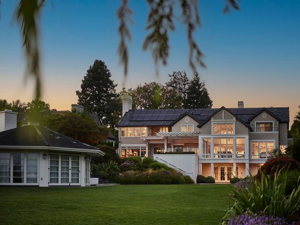 A large, multi-storey residential house viewed from a landscaped lawn at dusk. The home has a symmetrical gabled roofline, dark roof tiles, light grey exterior walls, white trim, extensive illuminated windows, balconies, and a prominent glazed central section. A separate smaller pavilion-like structure sits to the left, surrounded by mature trees, shrubs, ornamental grasses, and garden planting.