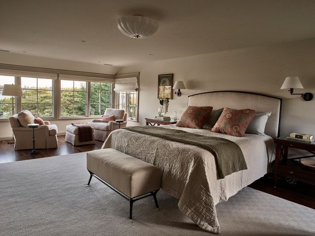 A spacious, refined bedroom with a large upholstered bed, textured neutral bedding, patterned accent cushions, and a matching upholstered bench. The room includes dark timber bedside tables, wall-mounted lamps, a framed artwork, and a sitting area by broad corner windows with two armchairs and a small side table. Soft natural light, neutral tones, and plush textiles create a calm, traditional atmosphere.