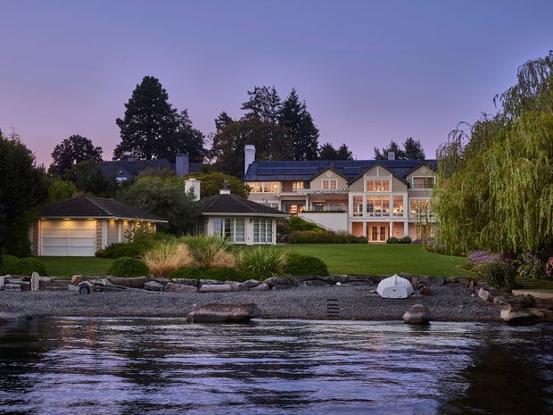 A twilight exterior view across calm waterfront toward a large multi-storey residential home with prominent gabled roof sections, pale rendered walls, extensive illuminated glazing, and attached structures. A manicured lawn, landscaped garden, gravel shoreline, rocks, and a small detached garage are visible in the foreground.