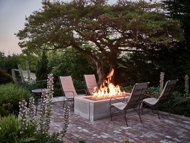 A landscaped outdoor patio with four lounge chairs arranged around a large square built-in fire pit. The patio is paved in weathered brick and surrounded by mature trees and garden planting, creating a secluded evening entertaining area.