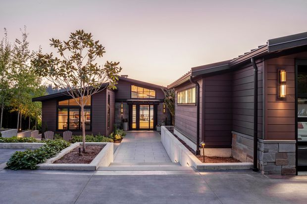 Exterior architectural view of a contemporary single-storey complex arranged around a paved entry courtyard. The buildings feature dark horizontal cladding, low-pitched rooflines, large glazed openings, stone-faced lower walls, integrated exterior lighting, and raised landscaped planters. The image is photographed at dusk with warm interior and facade lighting.