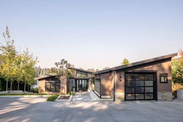 Exterior view of a low, contemporary single-storey home arranged around a paved driveway and entry path. The building has dark horizontal timber-style cladding, broad black-framed glazed garage doors and windows, stone-clad low walls, and asymmetrical mono-pitched roof forms. Mature landscaping and a spacious concrete forecourt frame the facade.
