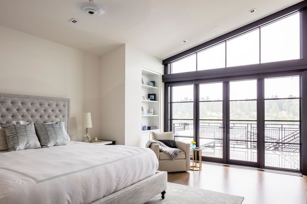 Bright, contemporary bedroom with a large upholstered tufted bed, layered neutral bedding, built-in open shelving, a compact armchair, and full-height dark-framed glazing opening to a balcony. The room has pale walls, light timber flooring, recessed ceiling lights, and a calm neutral palette.