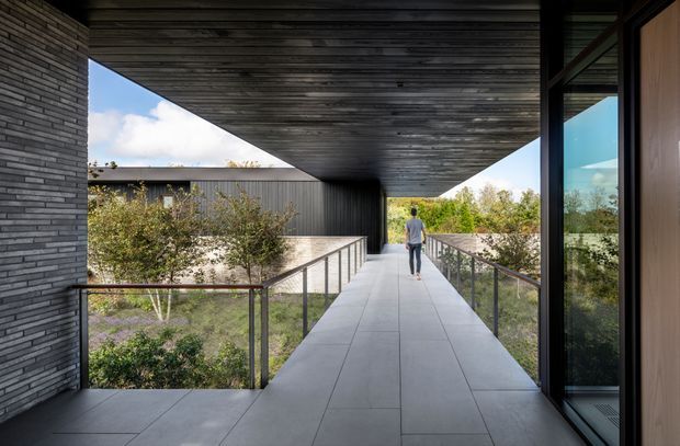 A covered exterior walkway or balcony-like passage connects modern building volumes. Large-format grey floor tiles extend beneath a dramatic dark timber-lined soffit, with slim dark metal and glass balustrades on both sides. A dark brick-clad wall is visible at left, while full-height glazing and warm timber cladding frame the right side. Landscaped gardens and a separate dark-clad structure are visible beyond, and a person provides scale.