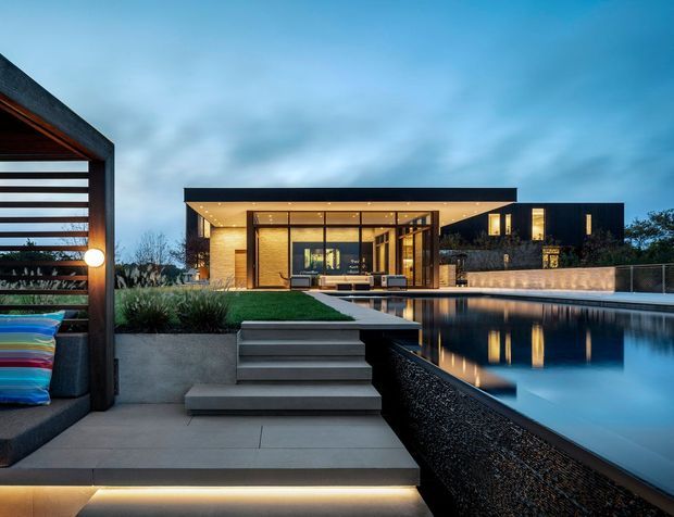 Dusk exterior view of a contemporary single-storey residence with a dark flat roof, extensive floor-to-ceiling glazing, warm timber-lined interior soffits, and a large reflecting swimming pool. A paved terrace with integrated step lighting, raised planters, and a slatted outdoor lounge structure occupies the foreground.