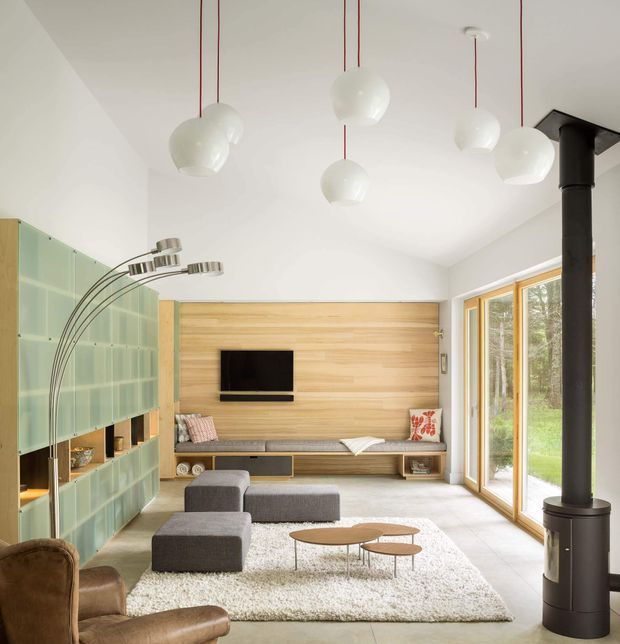 Bright contemporary living room with a built-in timber media wall and bench seating, modular grey upholstered seating, a large pale rug, sculptural pendant lights, and a tall black freestanding wood-burning stove. A translucent green built-in shelving wall occupies the left side, while full-height timber-framed glazing opens toward a leafy exterior.