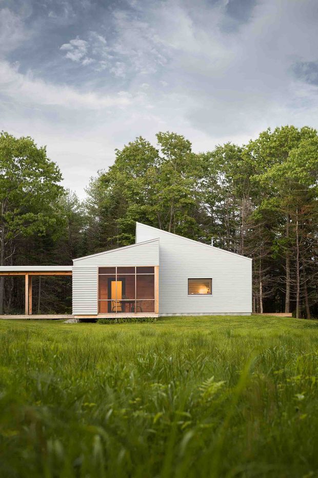 A small modern single-storey house set in a grassy clearing within a dense wooded landscape. The building has a simple asymmetrical pitched roof, pale horizontal metal or weatherboard-like cladding, extensive glazed doors at the front, and a covered open-sided extension or deck to the left. Warm interior light is visible through the windows beneath a dramatic overcast sky.