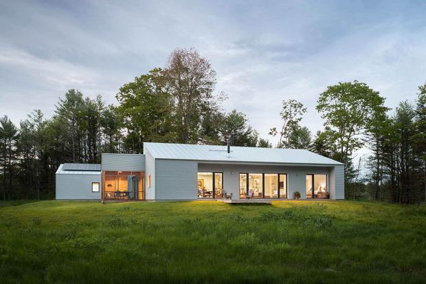 A single-storey contemporary house set within a grassy clearing and surrounded by mature woodland. The building has a long, low rectangular form with pale grey horizontal cladding, a shallow pitched metal roof, extensive floor-to-ceiling glazing along the main facade, and a smaller connected wing at the left. Warm interior lighting is visible through the windows, creating contrast with the cool evening exterior.