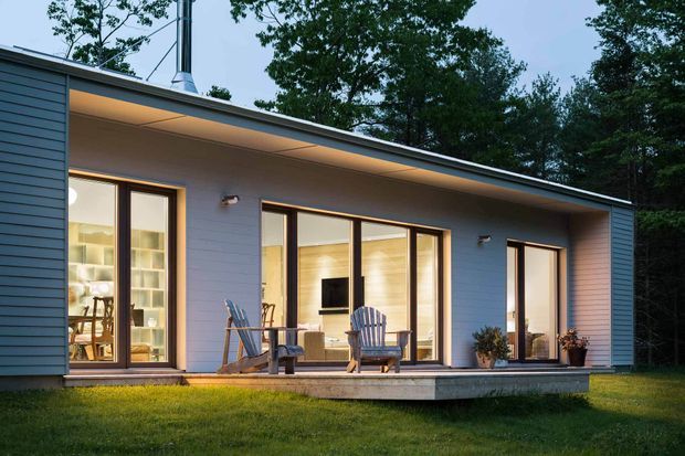 Single-storey contemporary house set among mature trees, with a long low roof, horizontal blue-grey cladding, broad glazed openings, and a covered timber deck. Warm interior lighting reveals living and dining areas, while outdoor lounge chairs and potted plants occupy the deck.