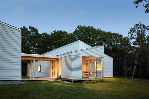 Exterior view of a contemporary single-storey house in a wooded setting. The building is composed of simple interconnected volumes with pale horizontal cladding, pitched gable roofs, large glazed openings, and a covered timber-framed outdoor walkway or porch. Warm interior lighting is visible through the windows and the house appears slightly elevated above a landscaped lawn.