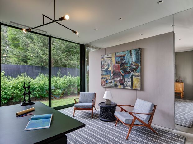 A refined contemporary living room or lounge with floor-to-ceiling corner glazing overlooking a planted garden. The room features a dark green rectangular table, two mid-century-inspired timber and upholstered lounge chairs, a small dark round side table, a large abstract artwork, a patterned area rug, and a linear multi-light pendant. A textured pale wall panel and concealed doorway lead toward an adjacent tiled space.