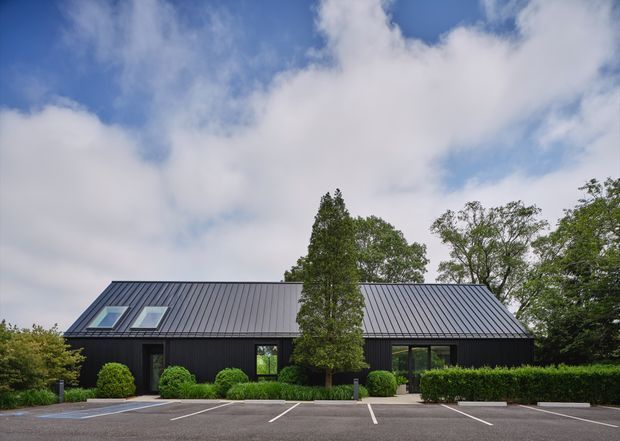Single-storey contemporary black-clad building with a steep pitched standing-seam metal roof, roof skylights, dark-framed windows, and neatly landscaped hedges and shrubs. The building fronts a marked paved parking area and is photographed from outside beneath a partly cloudy sky.