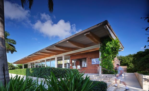 Exterior view of a low-slung contemporary house with broad projecting roof eaves, exposed timber soffits and beams, extensive floor-to-ceiling glazing, and warm horizontal timber cladding. Tropical landscaping, palms, dense hedging, and a paved access path surround the building.