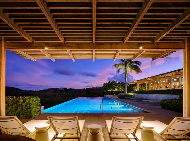 A dusk view from beneath a timber pergola or covered terrace, looking across a brightly illuminated swimming pool toward a low modern pavilion with extensive glazing. Lounge chairs and small side tables occupy the foreground, with tropical planting, a palm tree, and a hilly landscape beyond. The scene has warm timber overhead structure, cool blue pool lighting, and a dramatic purple-pink sunset sky.