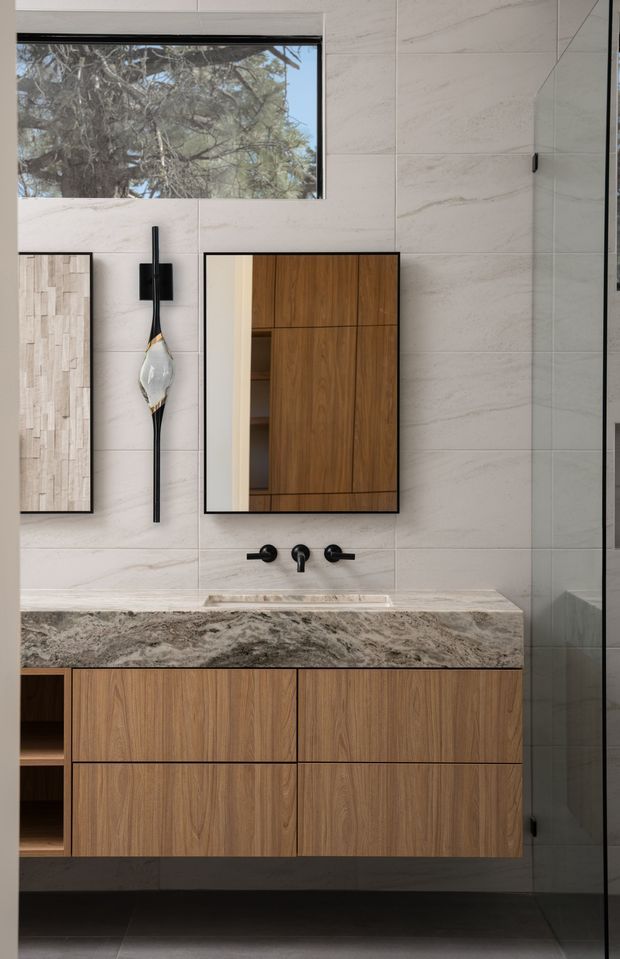 Contemporary bathroom vanity with a floating walnut-toned timber cabinet, heavily patterned natural stone benchtop, wall-mounted black tapware, large-format pale stone-look wall tiles, and a frameless glass shower enclosure. A horizontal window brings in natural light, while a rectangular mirror and sculptural wall sconce add refined detail.