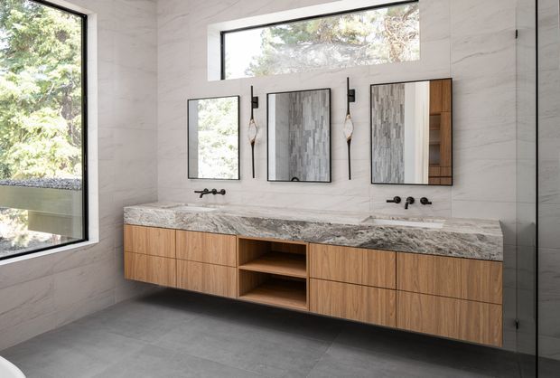 Contemporary double-vanity bathroom with a long floating timber vanity, open central shelving, pale veined stone benchtop, wall-mounted black tapware, and three mirrored cabinets. Large black-framed windows provide abundant natural light and views of greenery. The walls and floor are finished in large-format pale stone-look surfaces, creating a refined, warm modern interior.