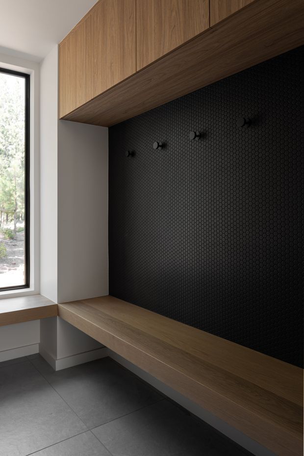 A contemporary built-in entryway or mudroom feature wall beside a tall window. The space has warm wood cabinetry and a matching floating bench, a dramatic black small-format hexagonal tile feature wall with dark hooks, white plastered side walls, and large dark grey floor tiles. The clean, integrated joinery creates a refined, functional storage and drop-zone area.