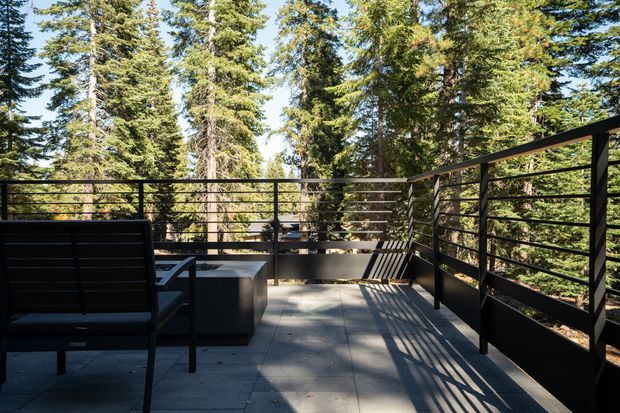 A contemporary outdoor terrace or deck surrounded by tall evergreen forest. The space has large square grey paving slabs, black metal horizontal-rail balustrades, a black outdoor lounge chair, and a dark rectangular side table. Strong sunlight creates linear shadows across the deck, giving the setting a private, natural and modern retreat atmosphere.