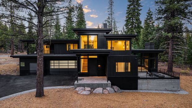 A contemporary multi-level dark-clad house set among tall forest trees. The facade features flat roof sections, extensive warm-lit glazing, a prominent central entry with an upper-level window, an integrated garage, and a raised concrete terrace with dark metal railings.
